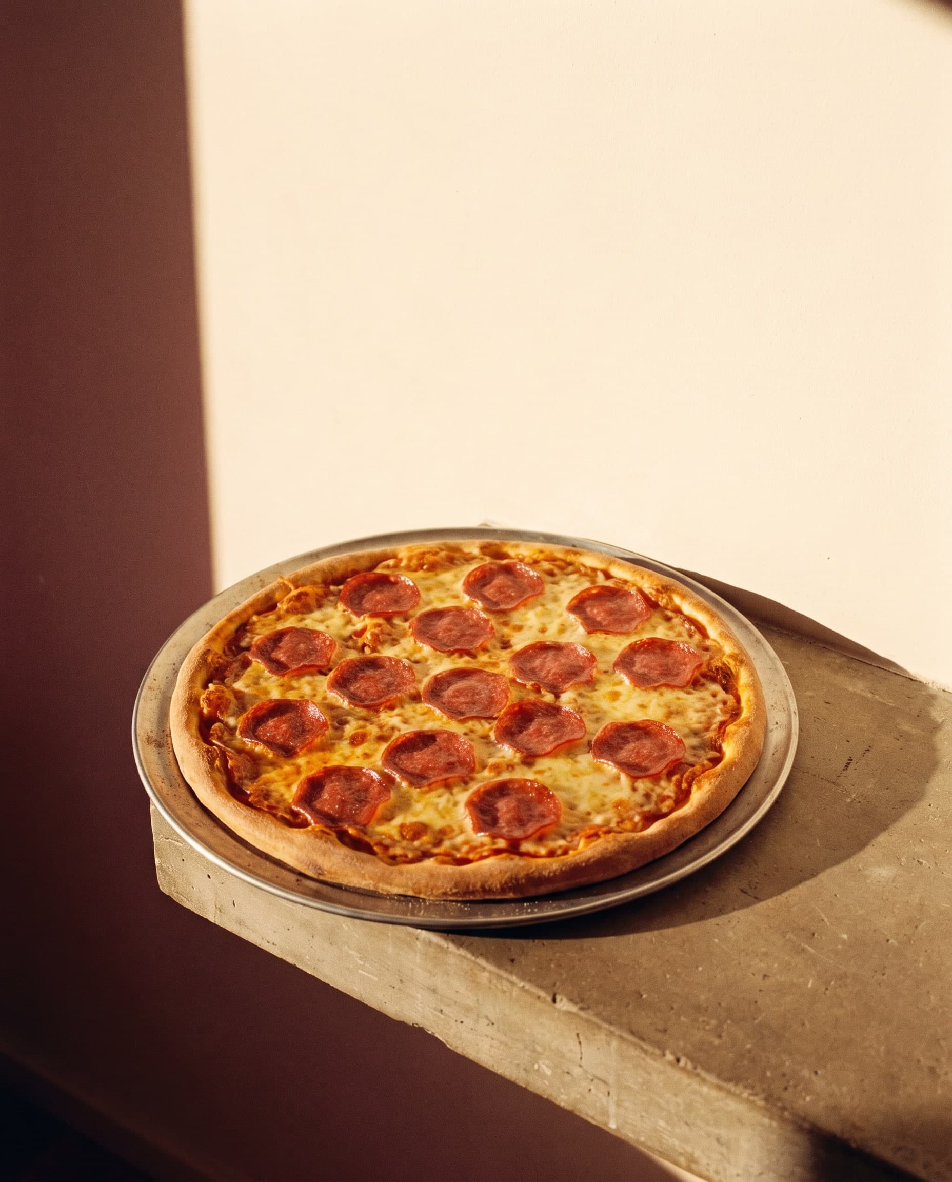 Salamipizza auf einem runden Blech vor einer hellen Wand im Streiflicht